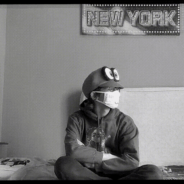a person wearing a mask and a hat sits on a bed in front of a sign that says new york
