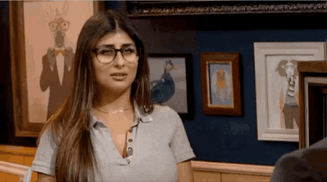 a woman wearing glasses and a gray shirt is standing in front of a wall with pictures on it .