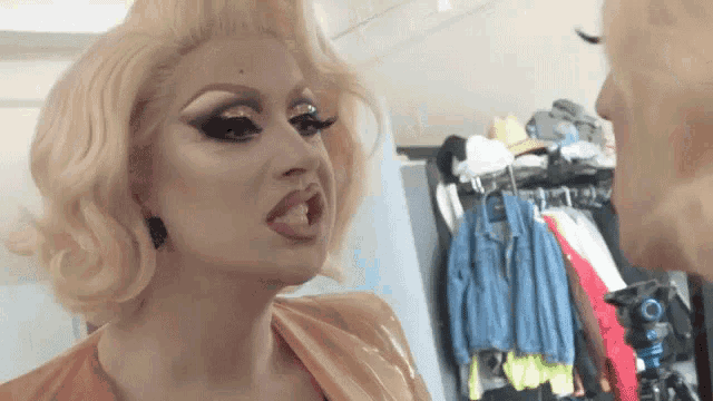 a drag queen is looking at another drag queen in front of a closet full of clothes