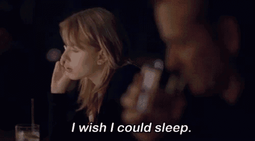 a woman sitting at a table with a glass of water and a man behind her saying " i wish i could sleep "