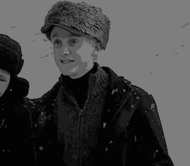 a black and white photo of a man wearing a fur hat standing next to a woman in the snow .