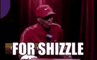 a man in a red hat is giving a speech and the words `` for shizzle '' are written on the screen behind him .