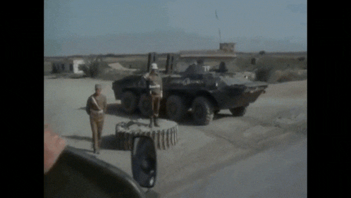 two soldiers stand in front of a military vehicle with the number 77 on the side