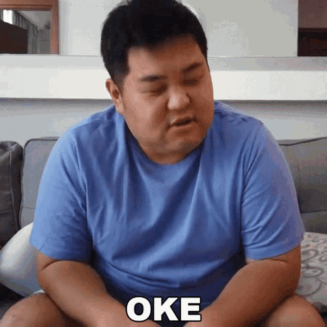a man in a blue shirt is sitting on a couch and the word oke is on his chest
