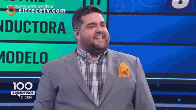 a man in a suit and plaid shirt stands in front of a screen that says 100 argentinas dicen
