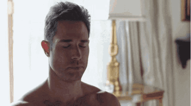 a shirtless man with his eyes closed is standing in front of a lamp in a room .