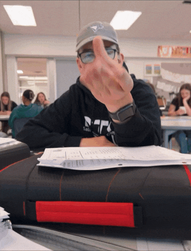 a man wearing a blue hat and glasses is giving the middle finger in a classroom