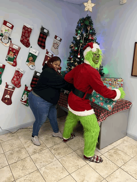 a woman stands next to a grinch in front of a christmas tree and stockings