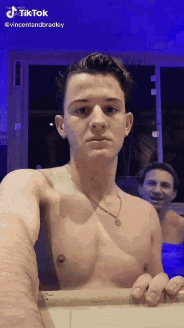 a shirtless man is taking a selfie in front of a mirror while another man looks on .