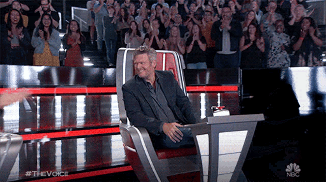 a man in a suit sits in a chair in front of a crowd that is clapping