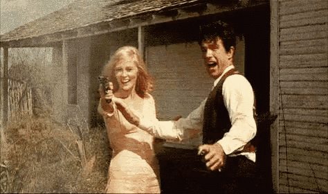 a man and a woman are standing in front of a building and the woman is pointing a gun at the man