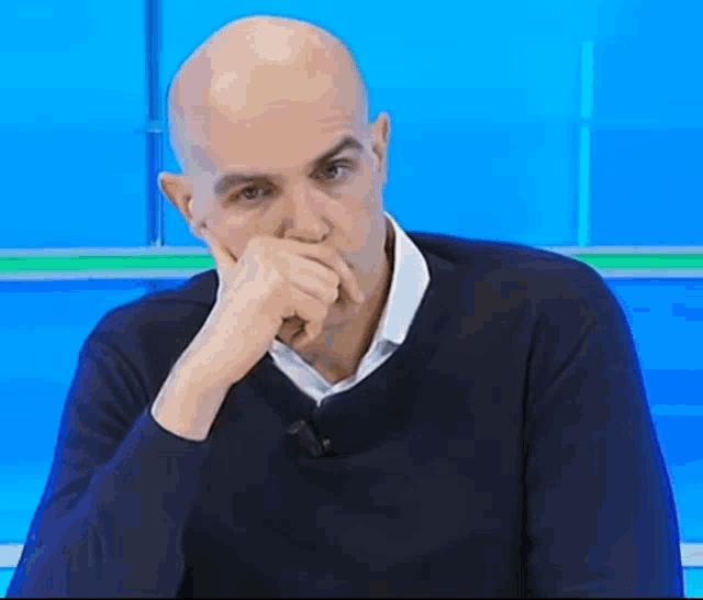 a bald man wearing a blue sweater covering his mouth with his hand