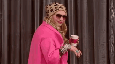 a woman in a pink coat and sunglasses is holding a cup .
