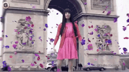 a woman in a pink dress is standing in front of an archway with purple flowers falling around her