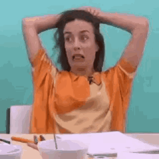 a woman in an orange shirt is sitting at a table with her hands on her head .