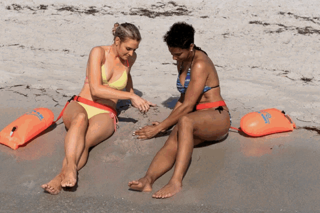 two women in bikinis are sitting on a beach with life preservers in the background