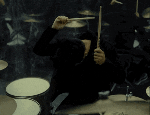 a man in a black hoodie plays drums in a dark room