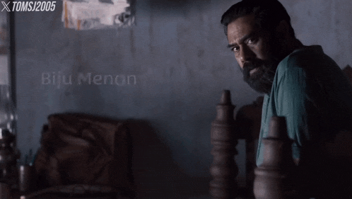 a man with a beard is sitting in a dark room with the name biju menon written on the bottom