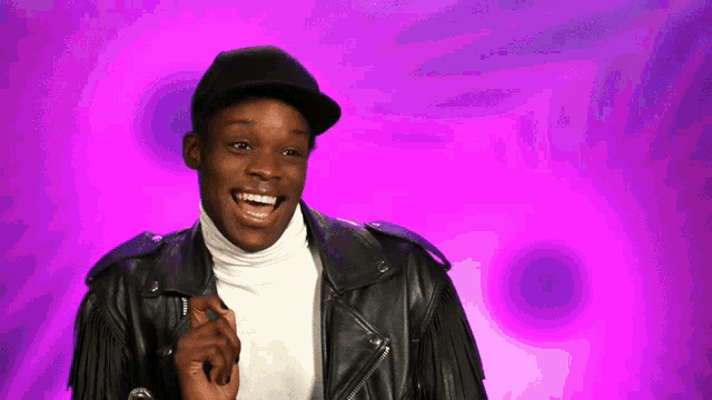 a man wearing a black hat and a leather jacket smiles in front of a purple background