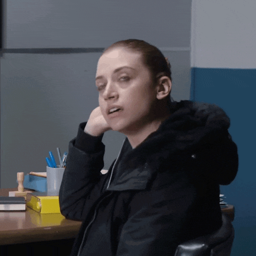 a woman in a black jacket sits at a desk with her hand on her chin