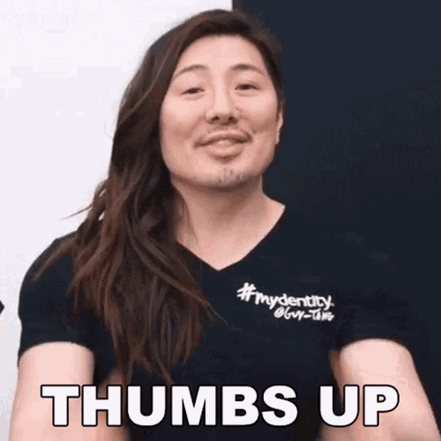a man with long hair and a beard is wearing a black shirt that says thumbs up