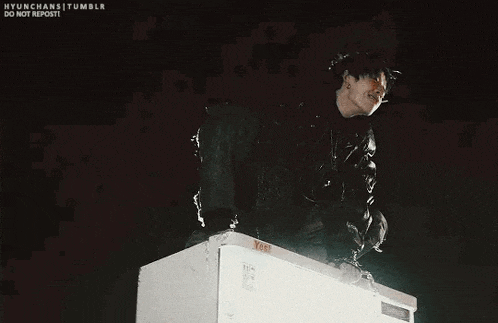 a man in a black jacket is standing on top of a white refrigerator .