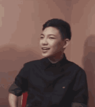 a young man wearing a black shirt with the name daebak on it is sitting in front of a red wall .
