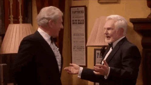 two men in tuxedos are talking to each other in a room with a lamp and a poster on the wall that says pantomimes