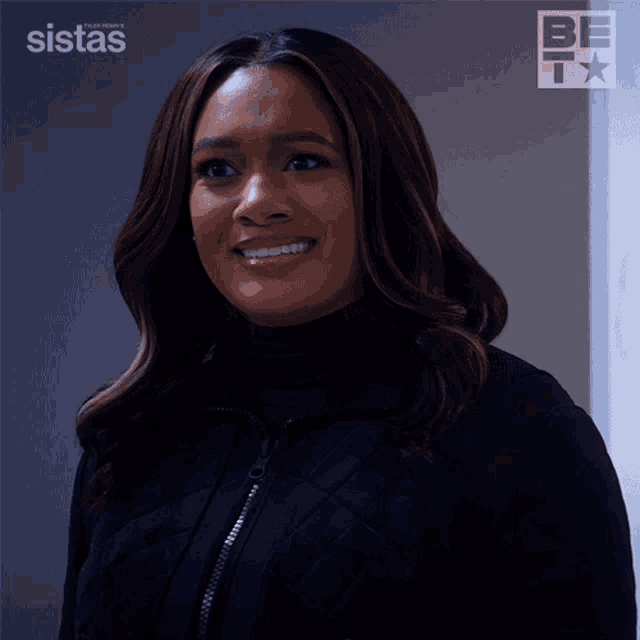 a woman in a black jacket is smiling in front of a sign that says sistas