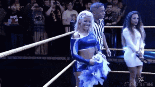 three women are standing in a wrestling ring with a referee and a crowd watching .