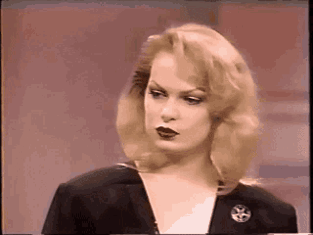 a woman with blonde hair and black lipstick is wearing a black jacket and a pin .
