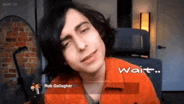 a young man with long hair is sitting in a chair with the words wait on the screen behind him