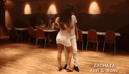 a man and a woman are dancing bachata in a room with tables and chairs