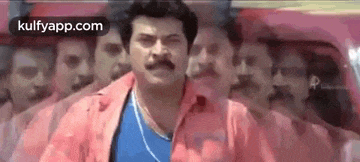 a man in a pink shirt and blue shirt is standing in front of a group of men .