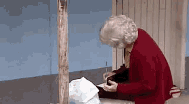 an elderly woman in a red sweater is sitting on the ground writing in a notebook .