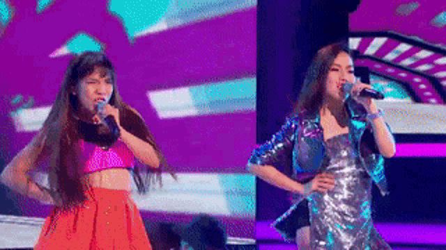 two women are singing into microphones on a stage in front of a purple background .