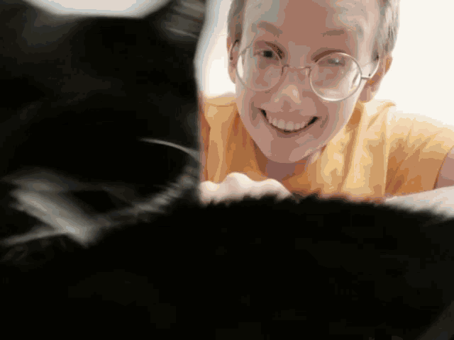 a man wearing glasses and a yellow shirt is smiling at a black cat