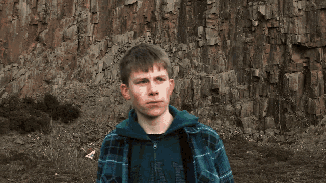 a young man in a plaid shirt stands in front of a large rock wall