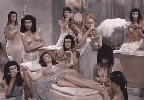 a group of women dressed in egyptian costumes are standing around a woman laying on a bed .
