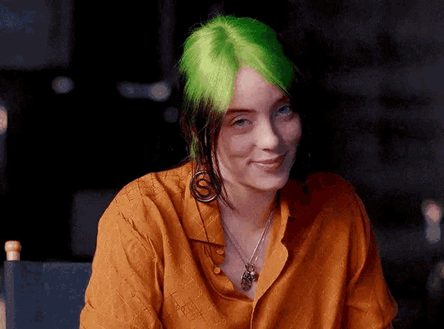 billie eilish has green hair and is wearing an orange shirt and earrings .