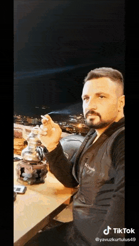a man with a beard is smoking a cigarette in front of a city at night .