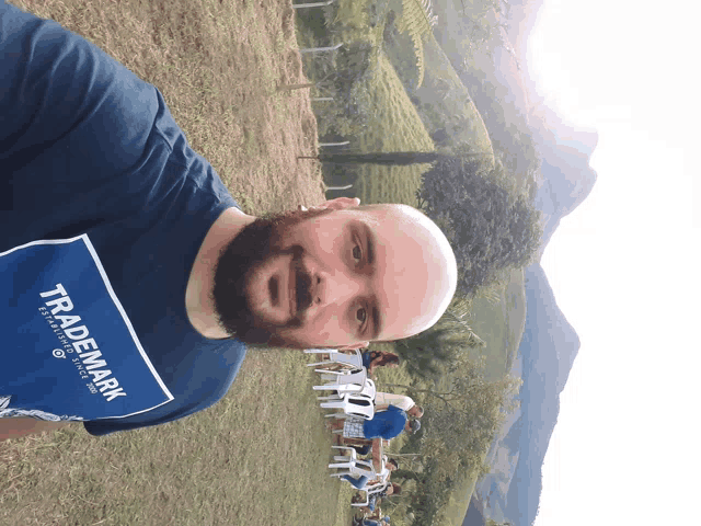 a bald man with a beard wears a blue shirt that says trademark