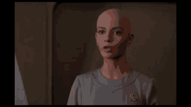 a woman with a bald head is wearing a grey shirt with an a on it