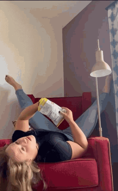 a woman is laying on a chair reading a book and holding a bag of chips