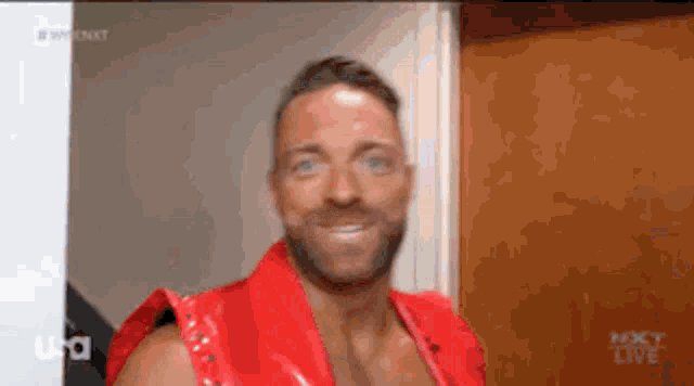 a man in a red vest is smiling in front of a tv screen that says nxt live