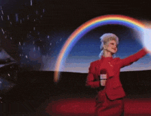 a woman in a red suit is standing in front of a rainbow and holding a microphone