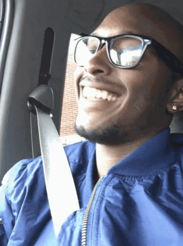 a man wearing glasses and a blue jacket is smiling while sitting in a car