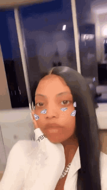 a woman with long black hair is wearing a white shirt and taking a selfie with diamonds on her face .