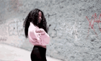 a woman in a pink sweatshirt is standing in front of a wall that has graffiti on it