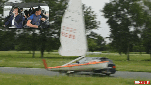 a car with a sailboat attached to it is driving down a road with trees in the background